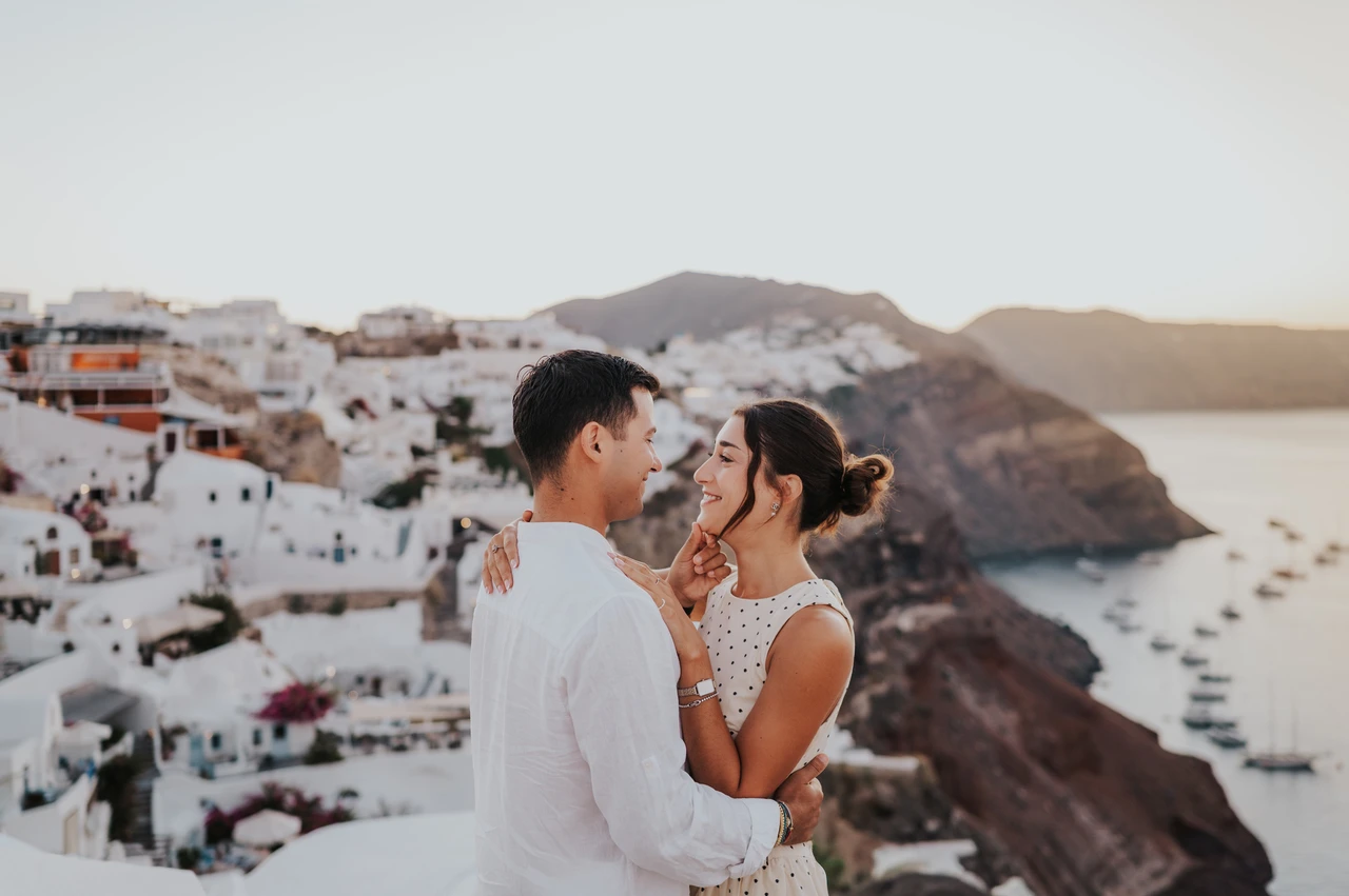 Santorini couple photoshoot: warm portrait above the caldera Couple standing close in warm light beside a white wall overlooking the caldera during their Santorini couple photoshoot.