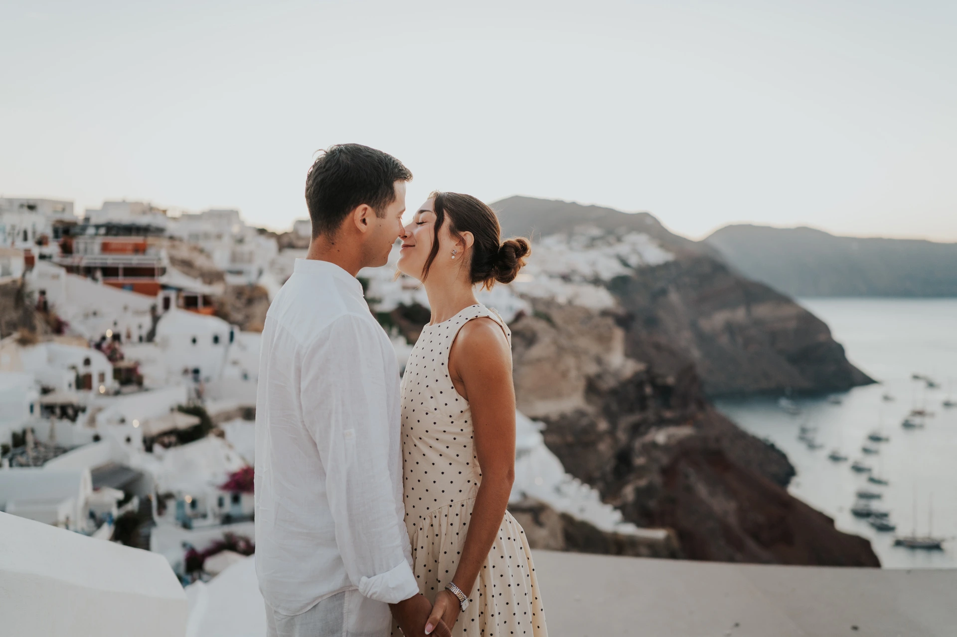 Couple kissing in a wide view with white buildings and the caldera stretching behind them during their Santorini couple photoshoot.