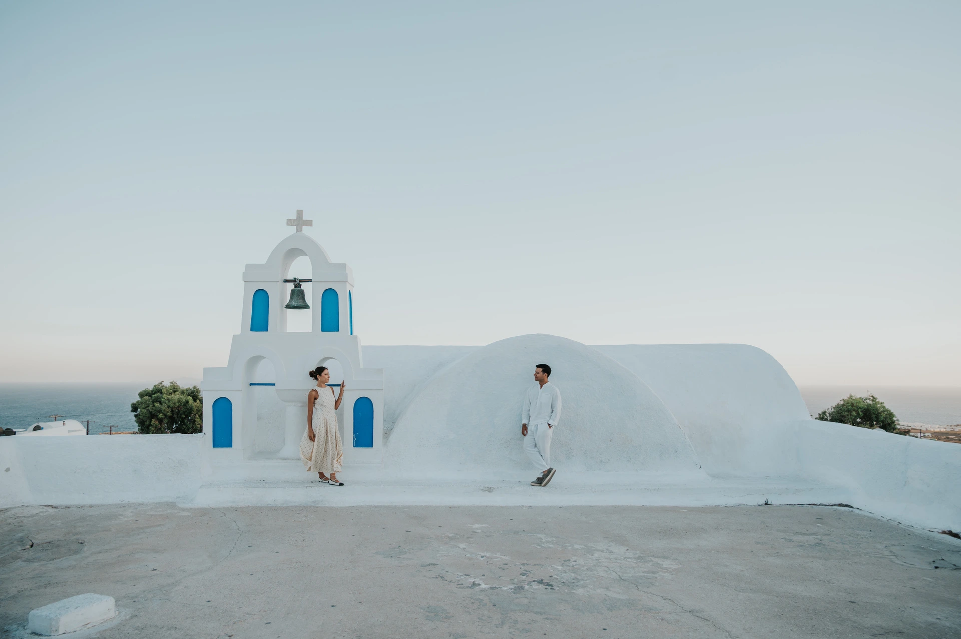 Wide view of a couple on a white terrace with a bell tower and the sea beyond during their Santorini couple photoshoot.