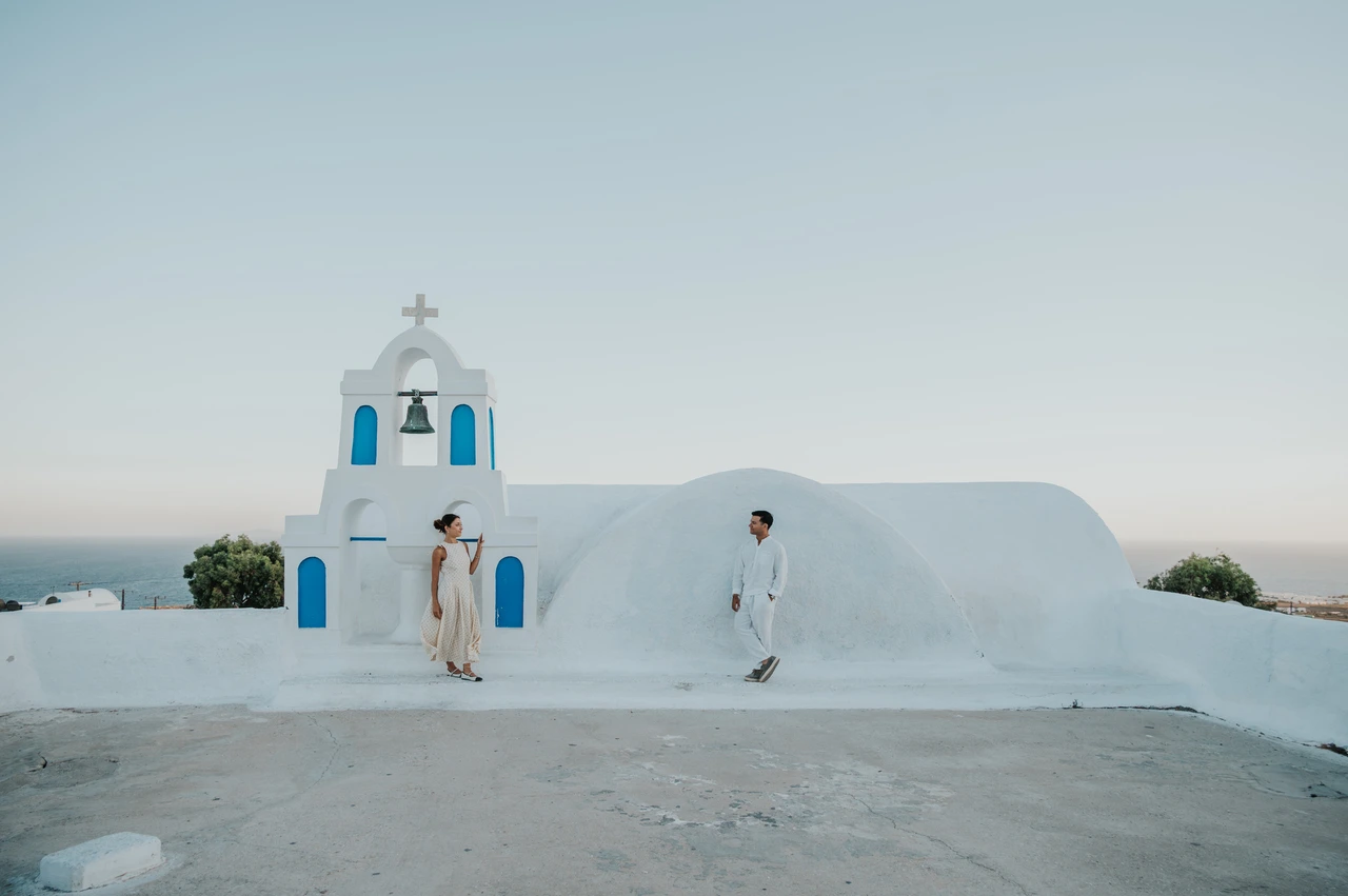Santorini couple photoshoot: wide portrait on a white bell-tower terrace Wide view of a couple on a white terrace with a bell tower and the sea beyond during their Santorini couple photoshoot.