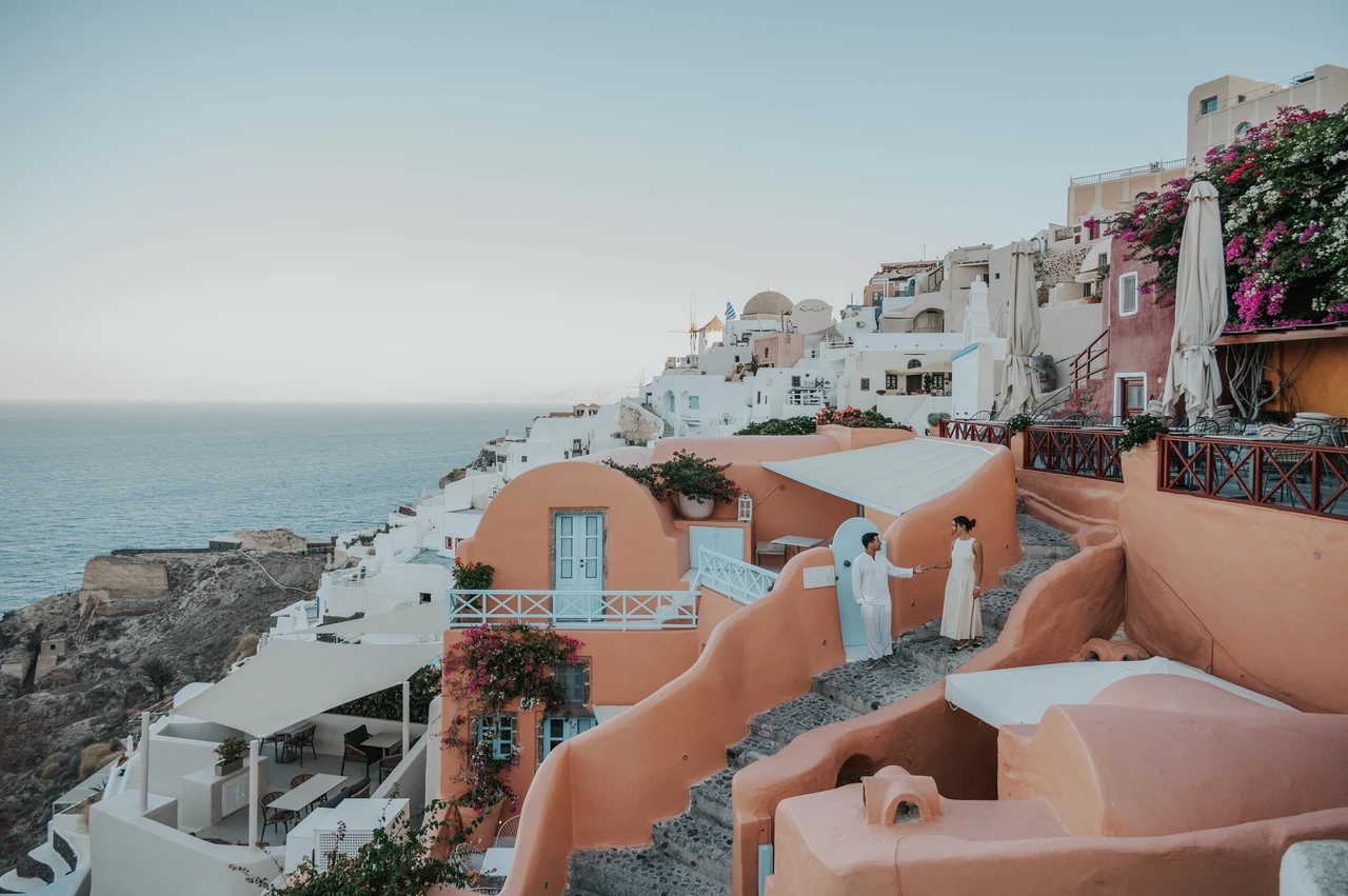 Santorini couple photoshoot: wide view of Oia’s winding stairs Wide view of a couple on winding steps between peach-coloured houses above the sea during their Santorini couple photoshoot.