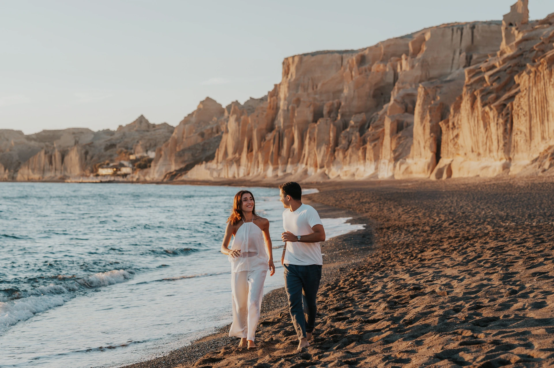 Couple walking together along dark sand beside the sea and pale cliffs during their Santorini couple photoshoot.