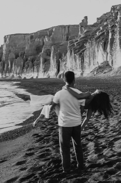 Black-and-white portrait of a man carrying his partner beside the sea below tall cliffs during their Santorini couple photoshoot.