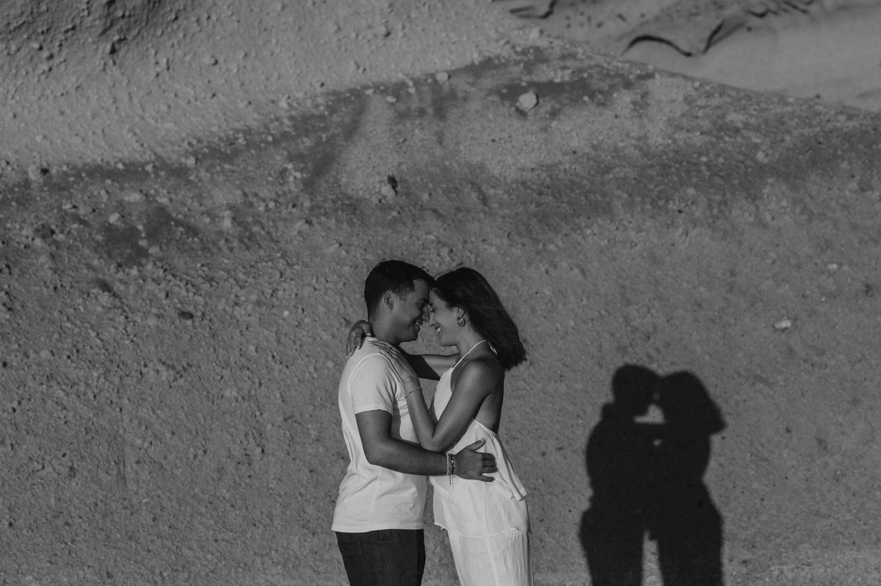Santorini couple photoshoot: black-and-white kiss against the cliffs Black-and-white portrait of a couple kissing below a rock wall with their shadow beside them during their Santorini couple photoshoot.