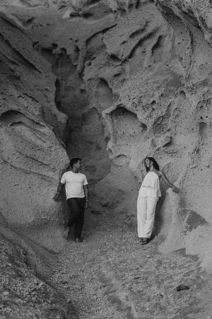 Santorini couple photoshoot: black-and-white portrait beside sculpted rock Black-and-white portrait of a couple standing on either side of a pale rock formation during their Santorini couple photoshoot.