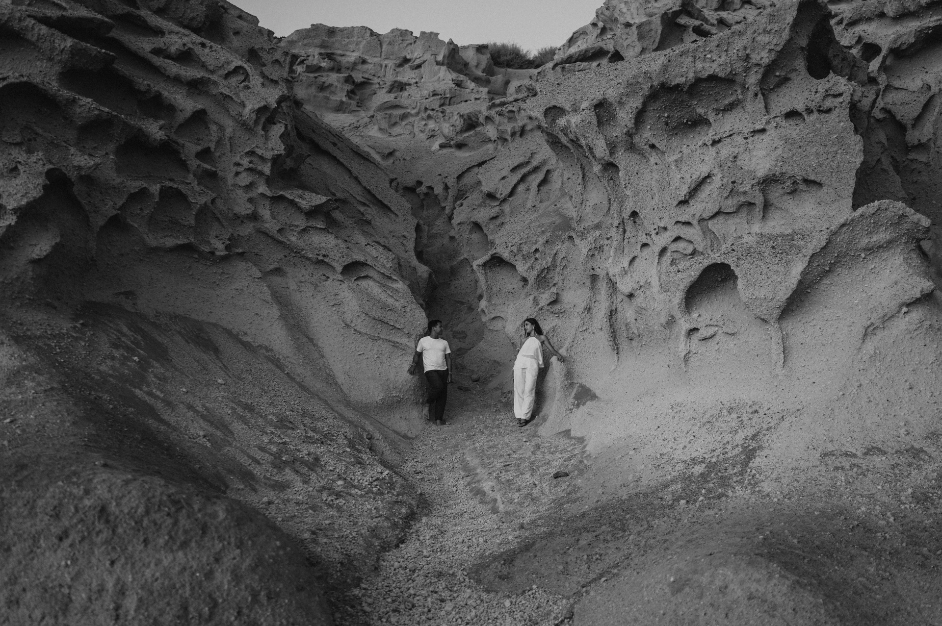 Black-and-white wide portrait of a couple standing in a sculpted rock alcove during their Santorini couple photoshoot.