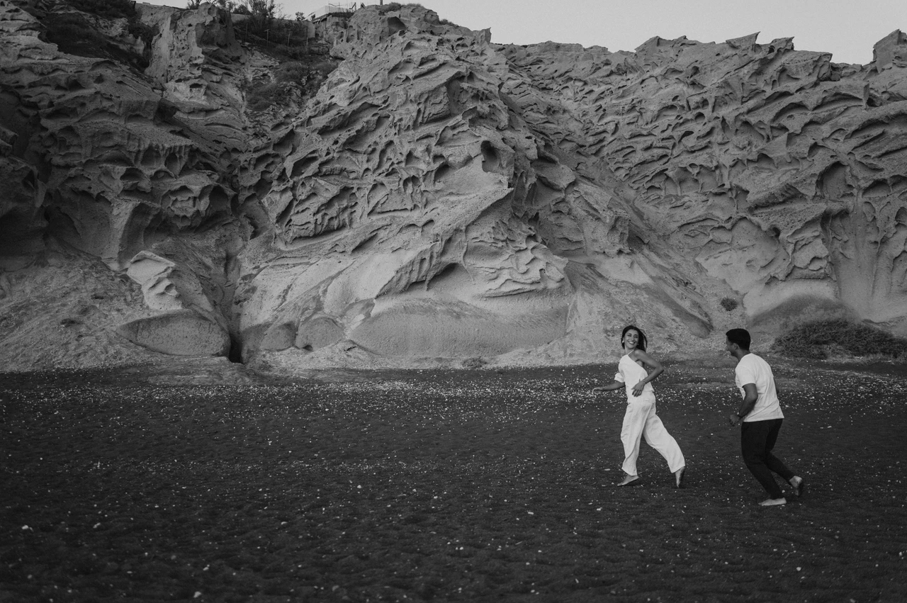 Santorini couple photoshoot: black-and-white run beneath the cliffs Black-and-white wide view of a couple running hand in hand below sculpted cliffs during their Santorini couple photoshoot.