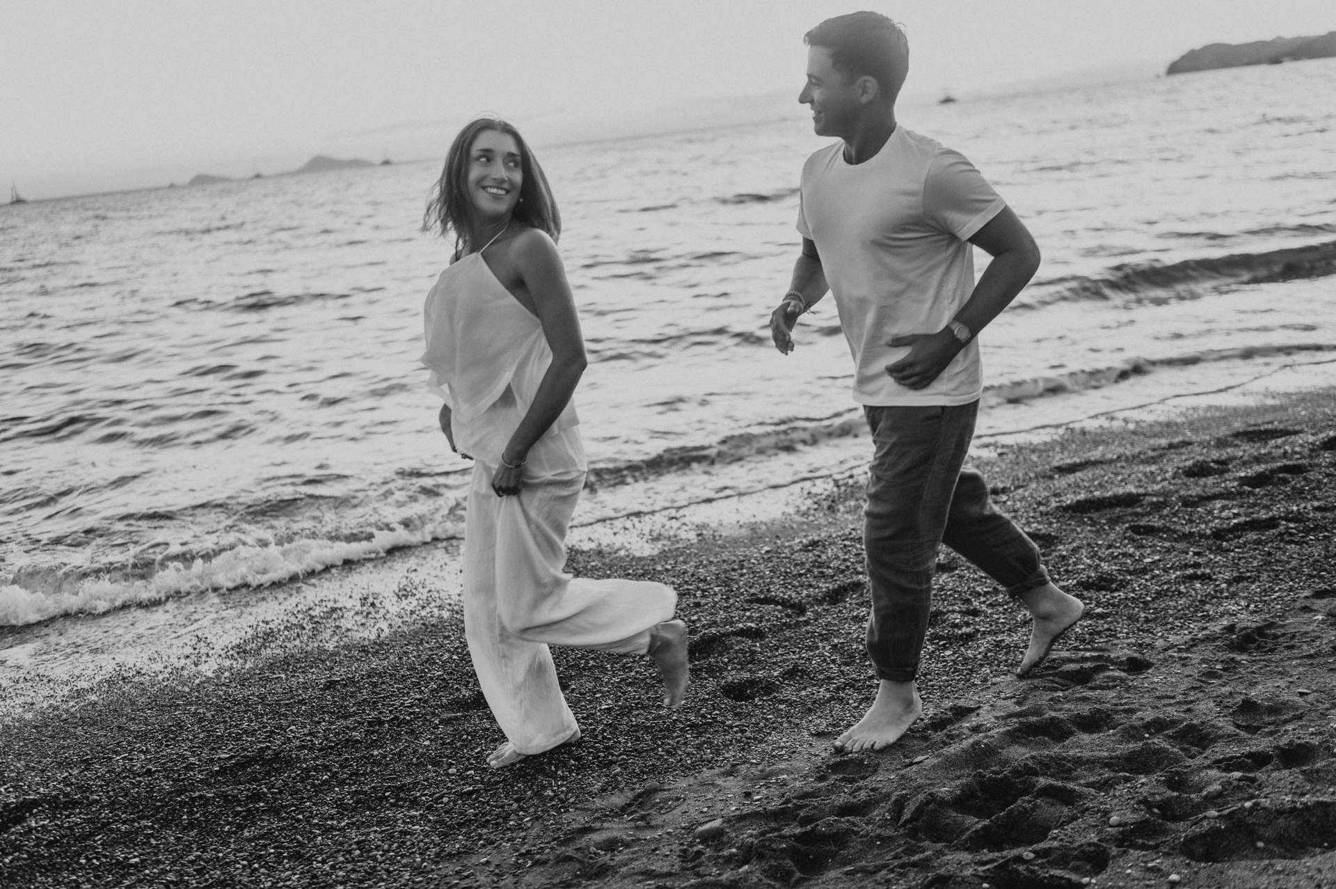 Black-and-white photograph of a couple running along the edge of the sea during their Santorini couple photoshoot.