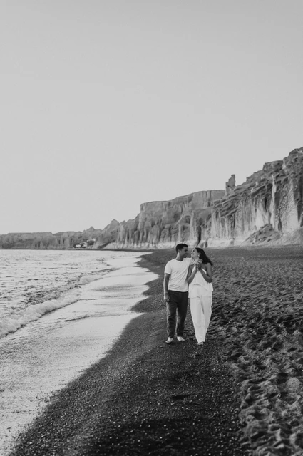 Santorini couple photoshoot: black-and-white shoreline portrait Black-and-white full-length portrait of a couple beside the sea beneath Vlychada’s cliffs during their Santorini couple photoshoot.