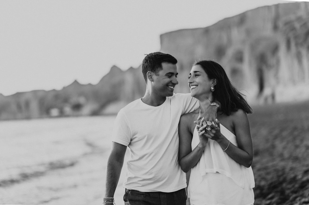 Santorini couple photoshoot: black-and-white smiles at Vlychada Beach Black-and-white portrait of a smiling couple with the sea and beach cliffs behind them during their Santorini couple photoshoot.