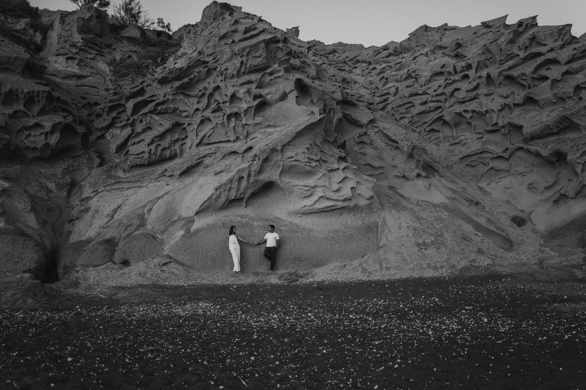 Black-and-white wide view of a couple standing beneath Vlychada’s sculpted cliff face during their Santorini couple photoshoot.