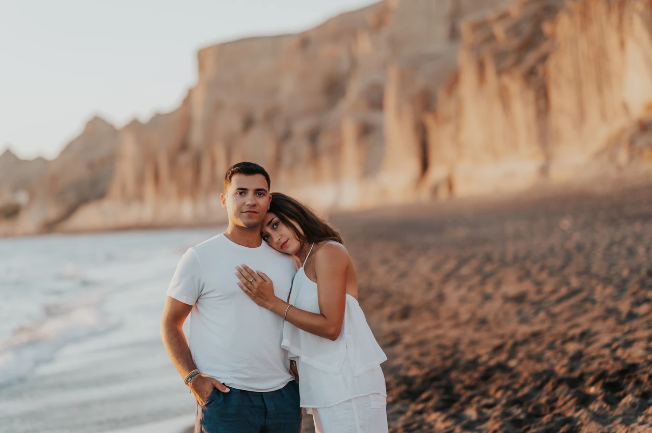Santorini couple photoshoot: close portrait on the dark-sand beach Couple standing close together on dark sand with pale cliffs softly blurred behind them during their Santorini couple photoshoot.