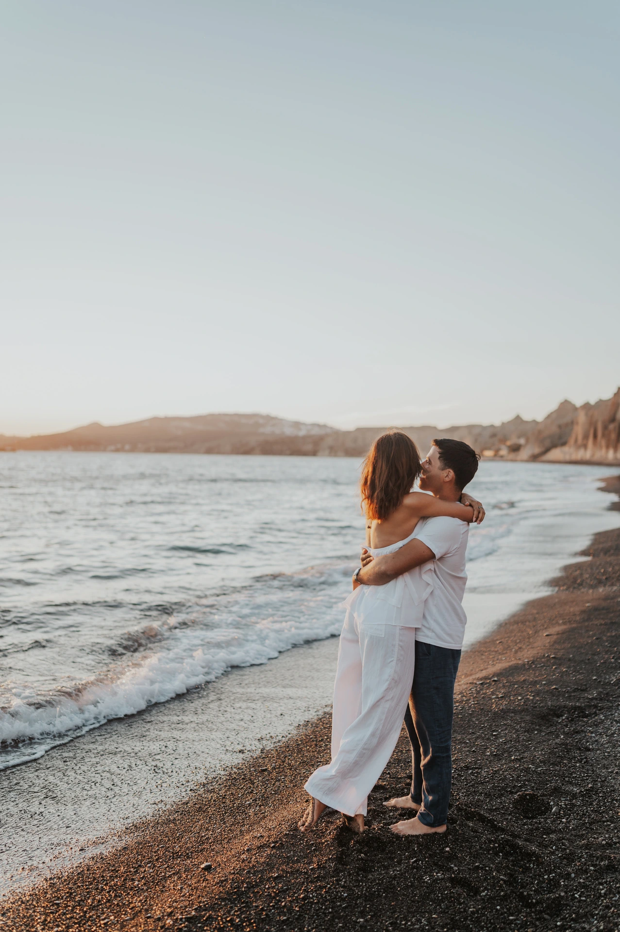 Couple embracing beside the water with Vlychada’s pale cliffs in the distance during their Santorini couple photoshoot.