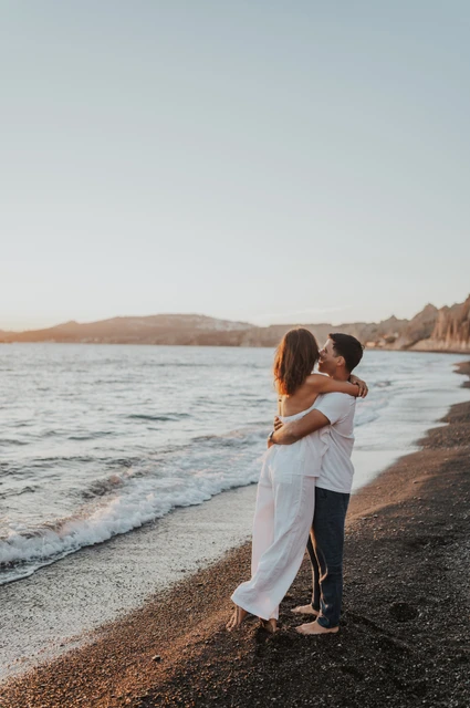 Santorini couple photoshoot: embrace beside Vlychada’s waterline Couple embracing beside the water with Vlychada’s pale cliffs in the distance during their Santorini couple photoshoot.