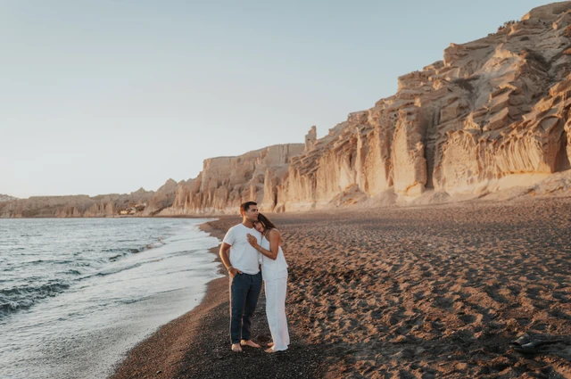 Wide view of a couple embracing on dark sand between the sea and pale cliffs during their Santorini couple photoshoot.