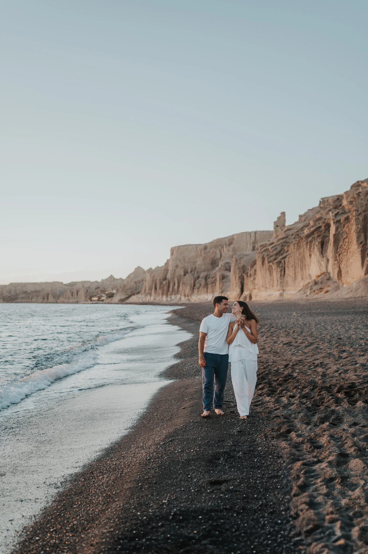 Full-length portrait of a couple standing together beside the sea below a long line of cliffs during their Santorini couple photoshoot.