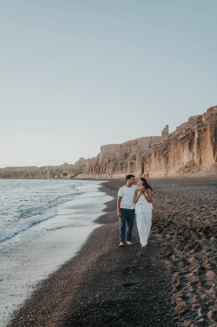 Santorini couple photoshoot: full-length portrait beside the shoreline Full-length portrait of a couple standing together beside the sea below a long line of cliffs during their Santorini couple photoshoot.