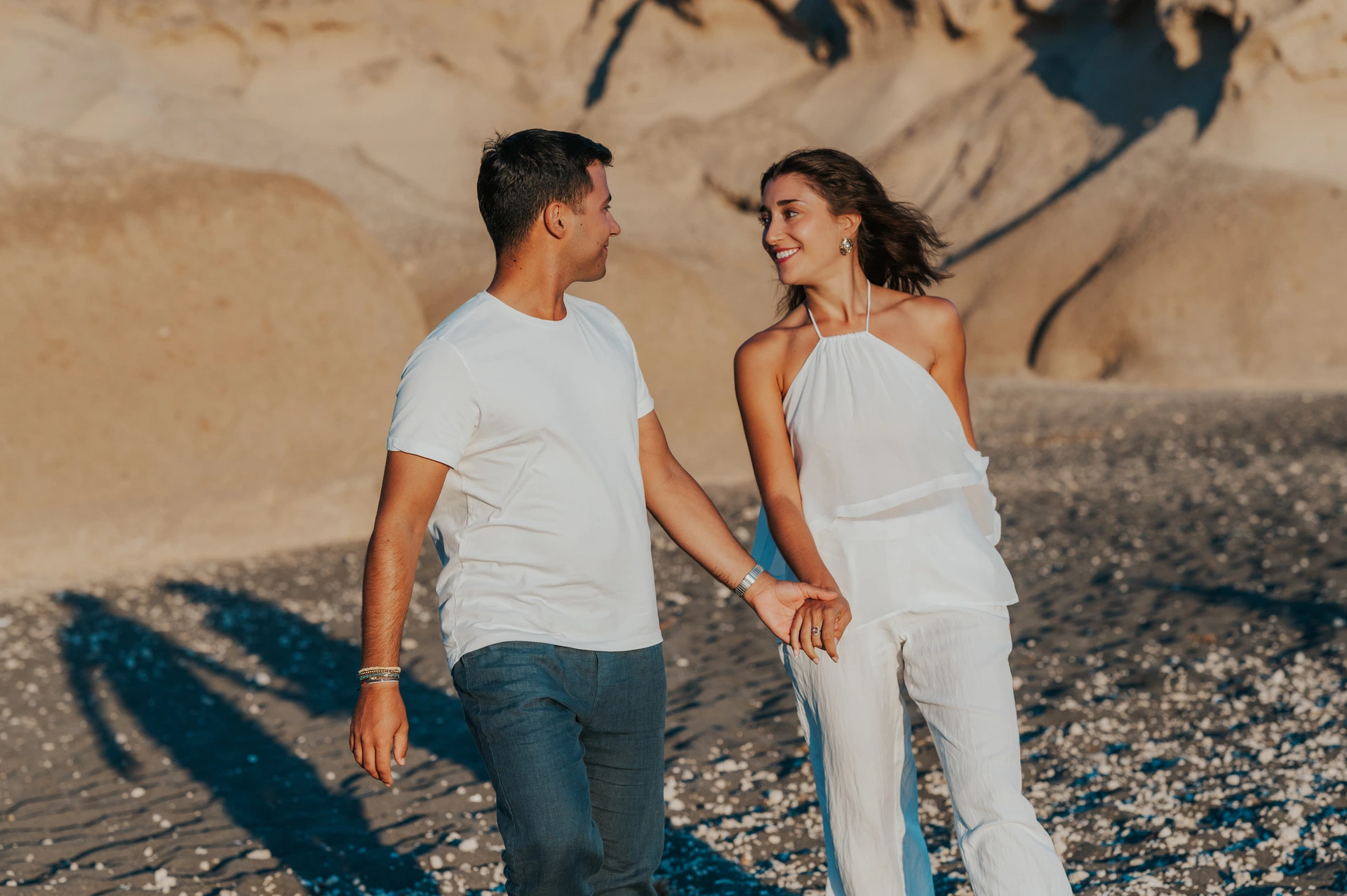 Couple holding hands and looking at one another on dark sand below pale sculpted cliffs during their Santorini couple photoshoot.
