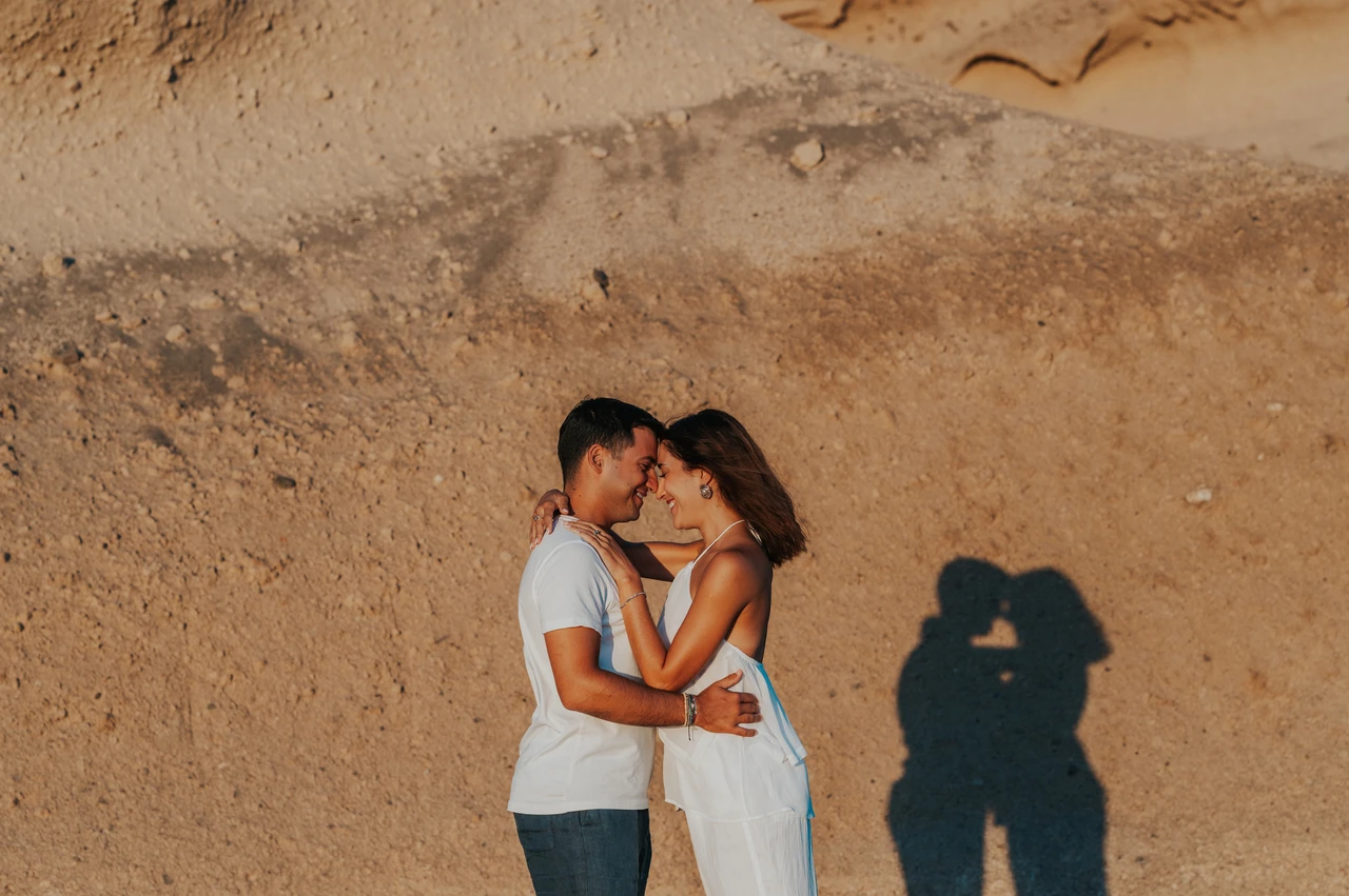 Santorini couple photoshoot: kiss and shadows against the beach cliffs Couple kissing below a sunlit rock wall with their shadow falling across the stone during their Santorini couple photoshoot.
