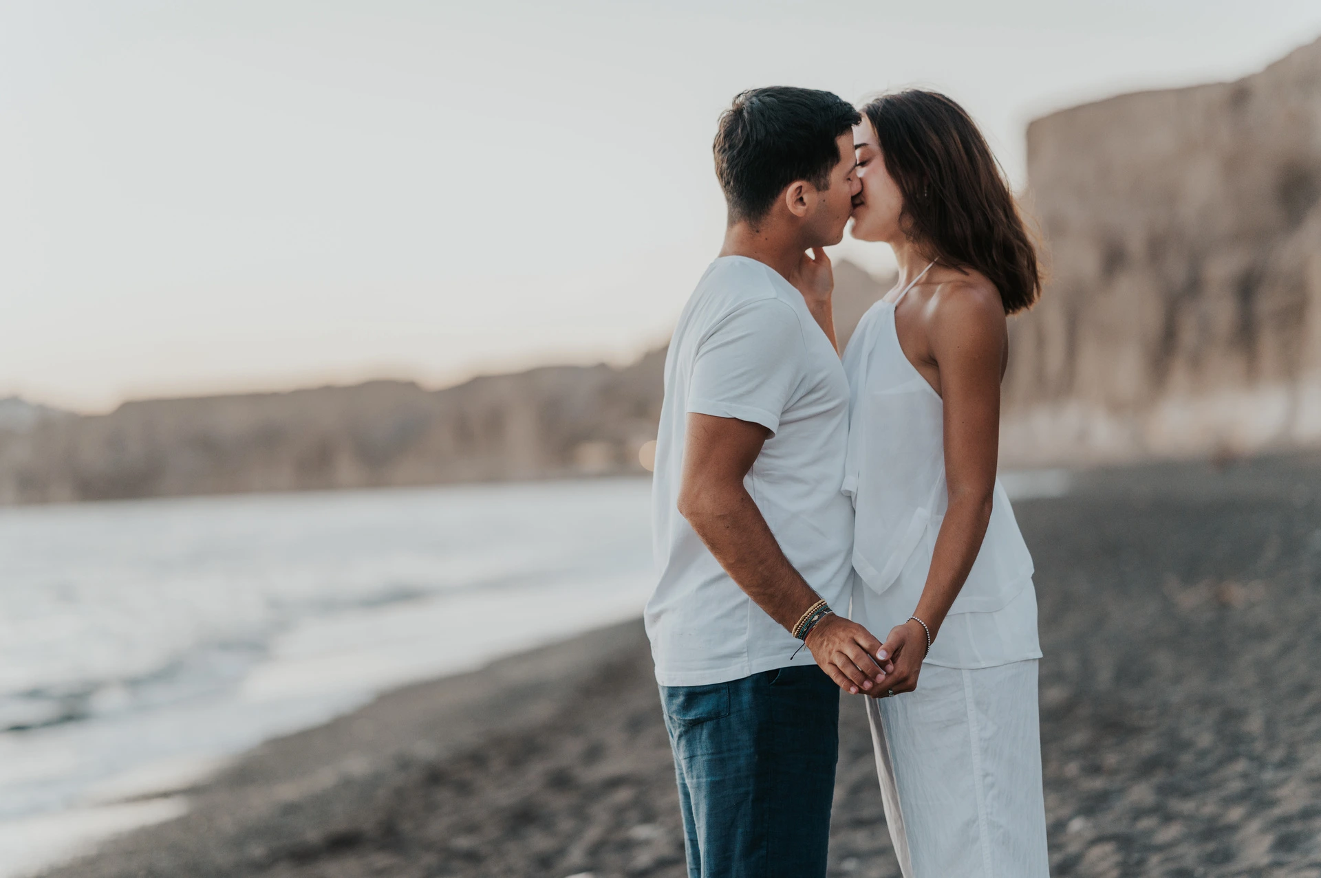 Couple kissing and holding hands beside the sea with cliffs softly blurred behind them during their Santorini couple photoshoot.