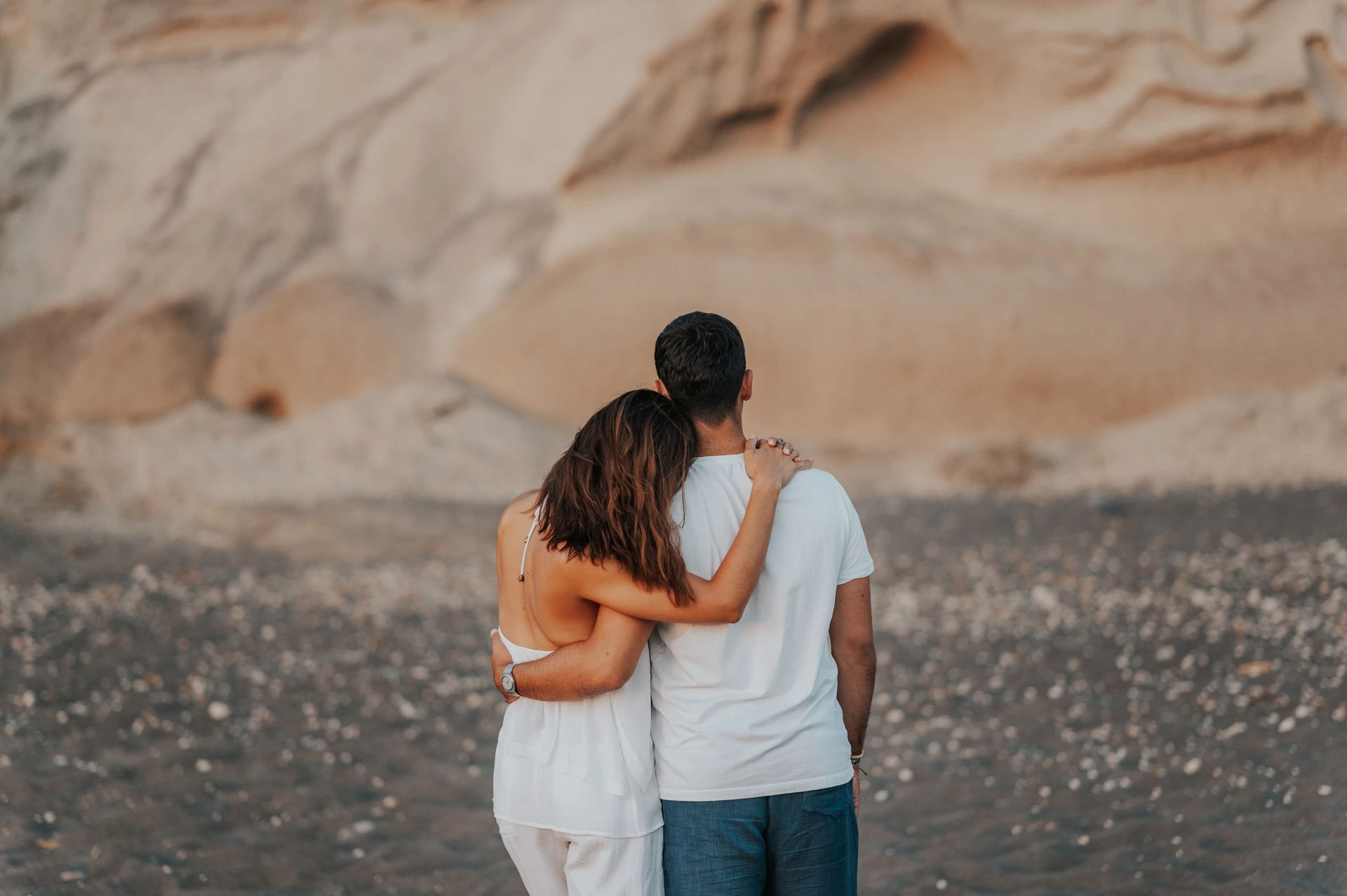 Couple seen from behind with arms around each other facing sculpted rock formations during their Santorini couple photoshoot.