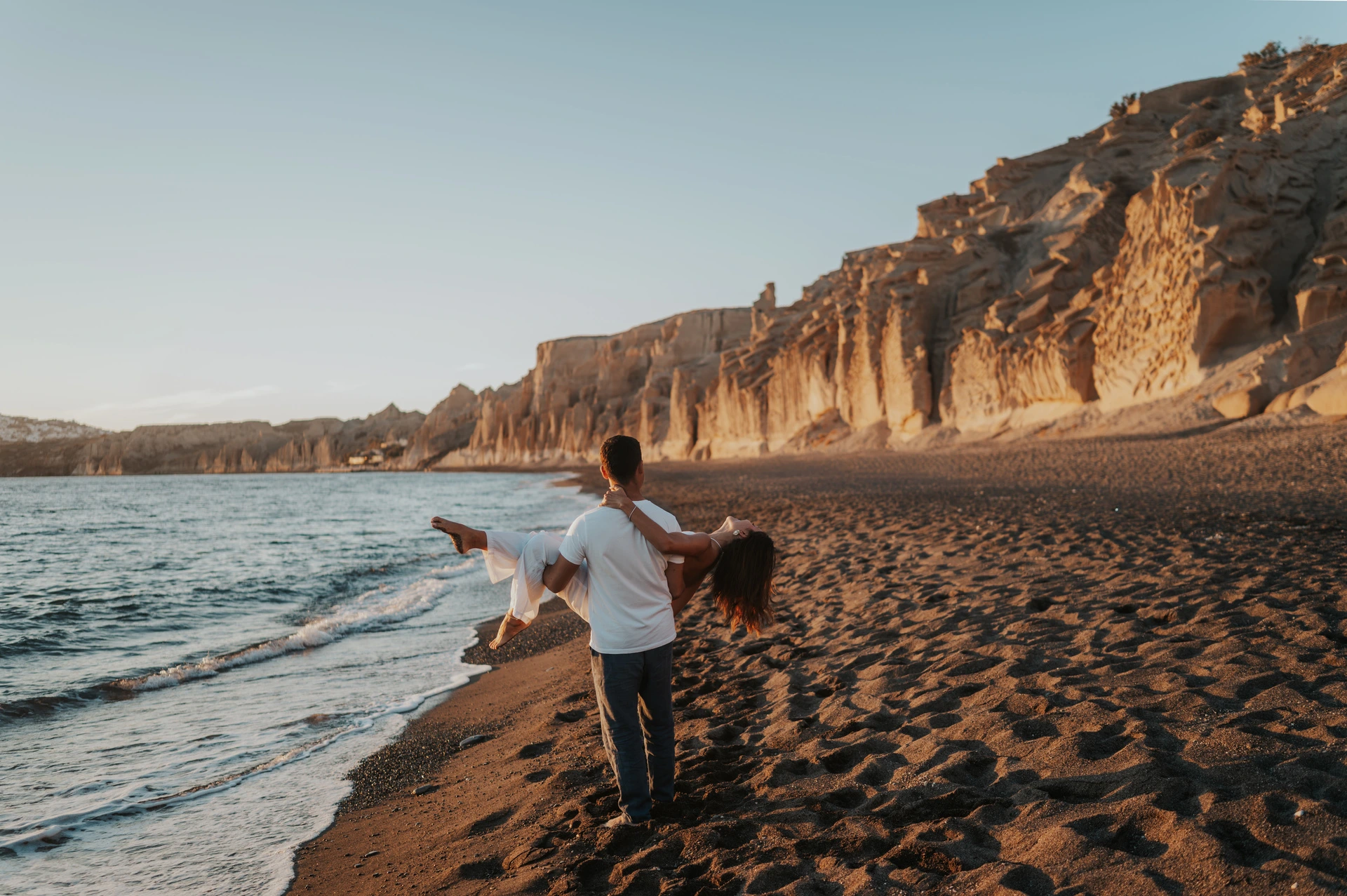 Man carrying his partner beside the water with Vlychada’s long cliff line behind them during their Santorini couple photoshoot.