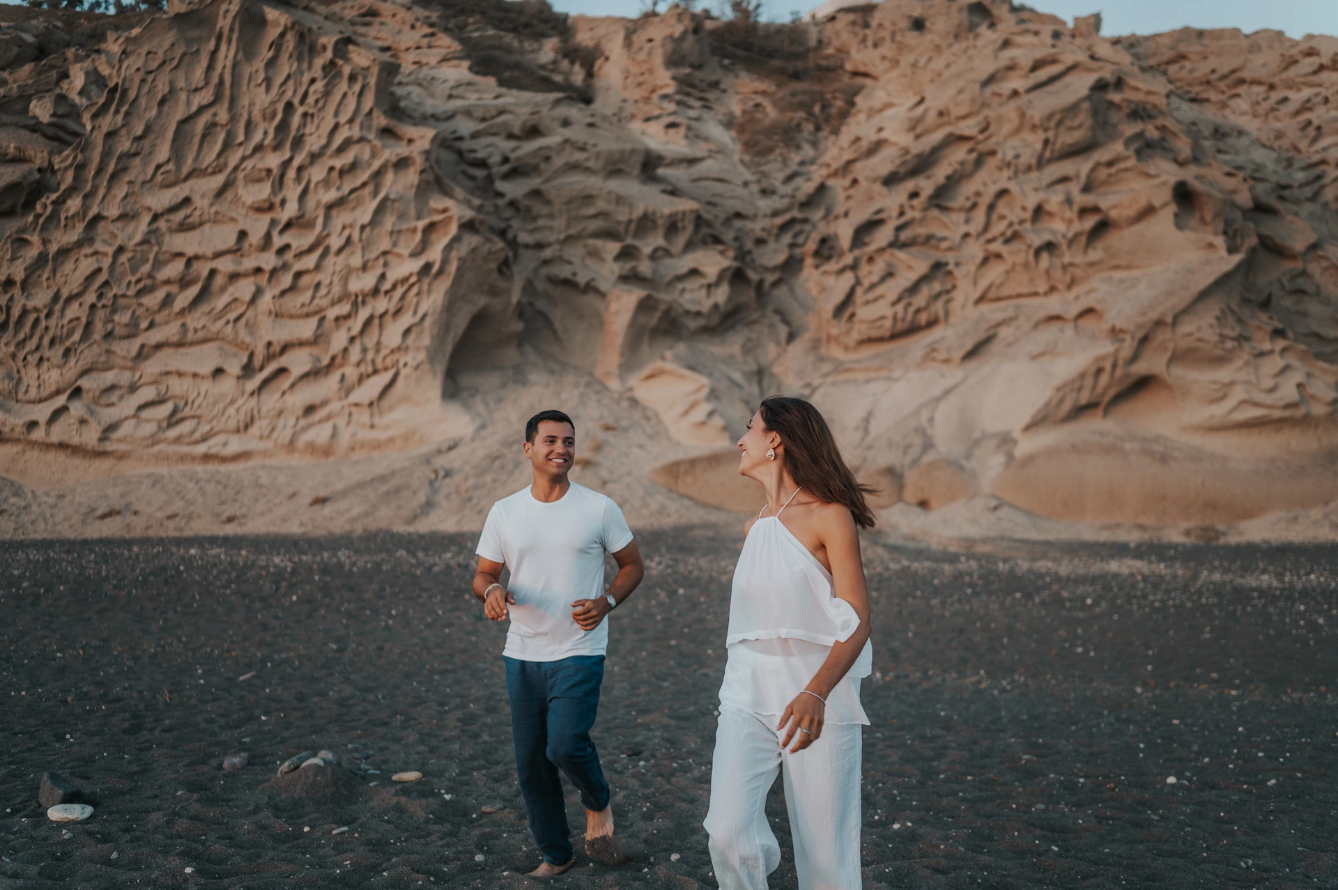Couple running together across dark sand with pale textured cliffs behind them during their Santorini couple photoshoot.