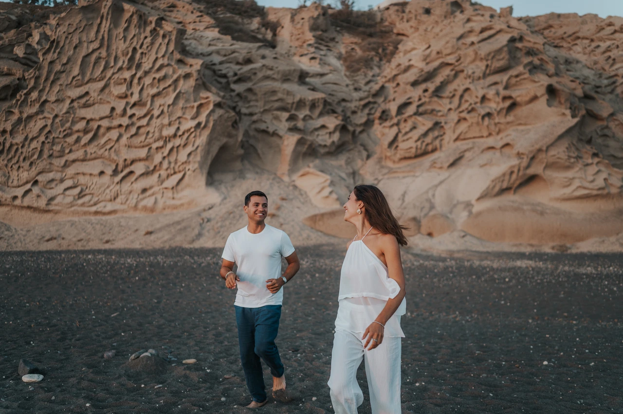 Santorini couple photoshoot: playful movement across the dark sand Couple running together across dark sand with pale textured cliffs behind them during their Santorini couple photoshoot.