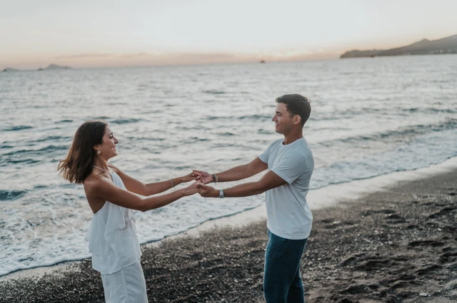 Couple holding both hands and leaning apart playfully beside the water during their Santorini couple photoshoot.