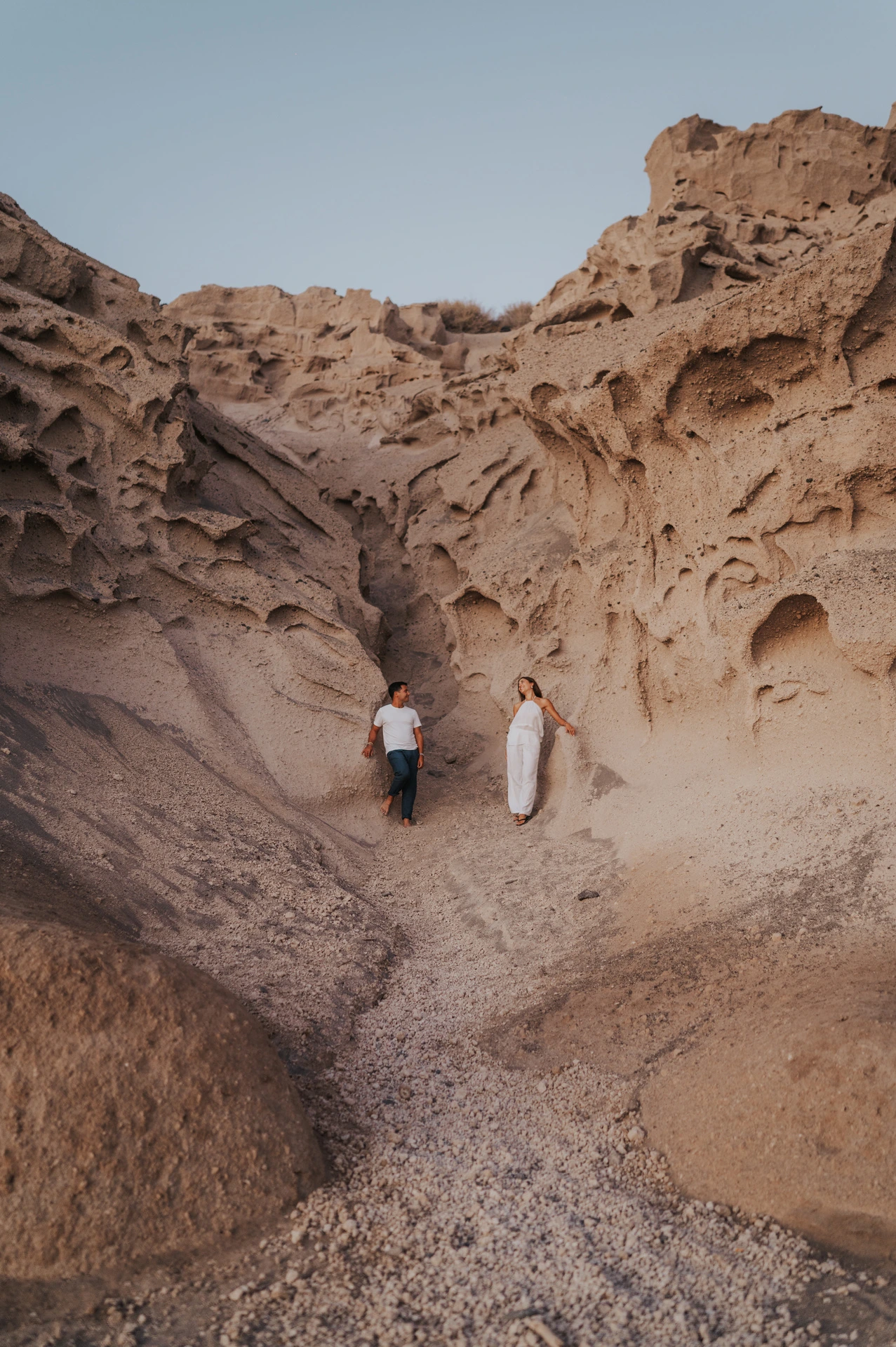 Full-length view of a couple beneath tall sculpted rock walls with dark sand below during their Santorini couple photoshoot.