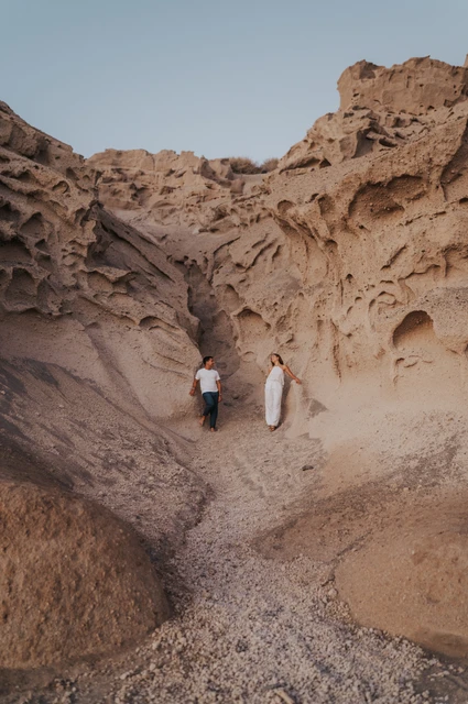 Full-length view of a couple beneath tall sculpted rock walls with dark sand below during their Santorini couple photoshoot.