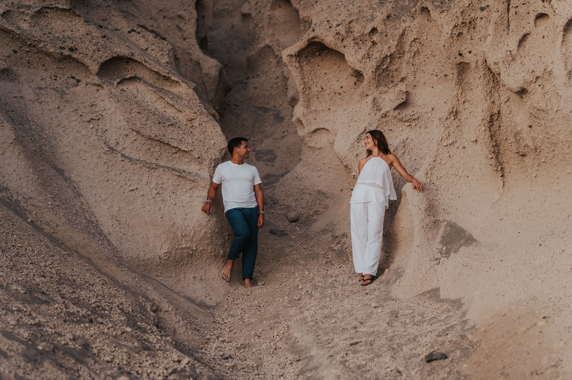 Couple standing on either side of a sculpted pale rock formation above the dark beach during their Santorini couple photoshoot.