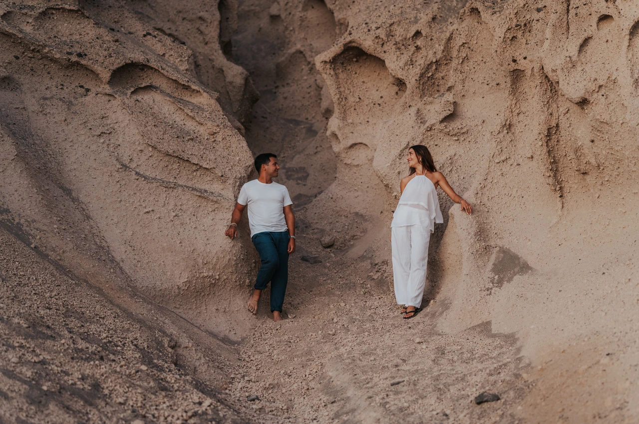 Santorini couple photoshoot: portrait beside Vlychada’s sculpted rock Couple standing on either side of a sculpted pale rock formation above the dark beach during their Santorini couple photoshoot.