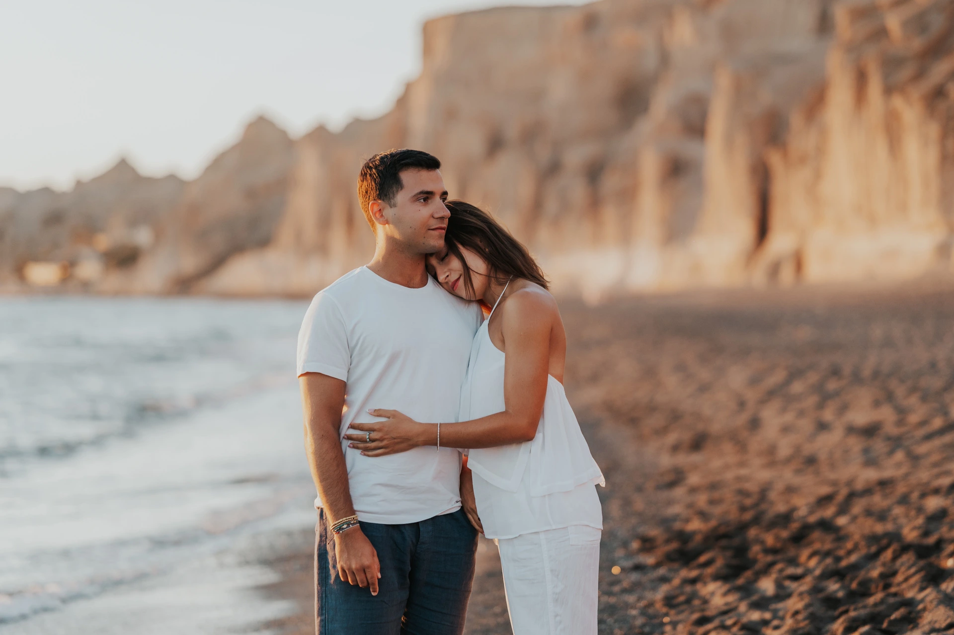 Santorini couple photoshoot: quiet portrait beside Vlychada’s cliffs Man kissing his partner’s head as she leans against his shoulder on the beach during their Santorini couple photoshoot.