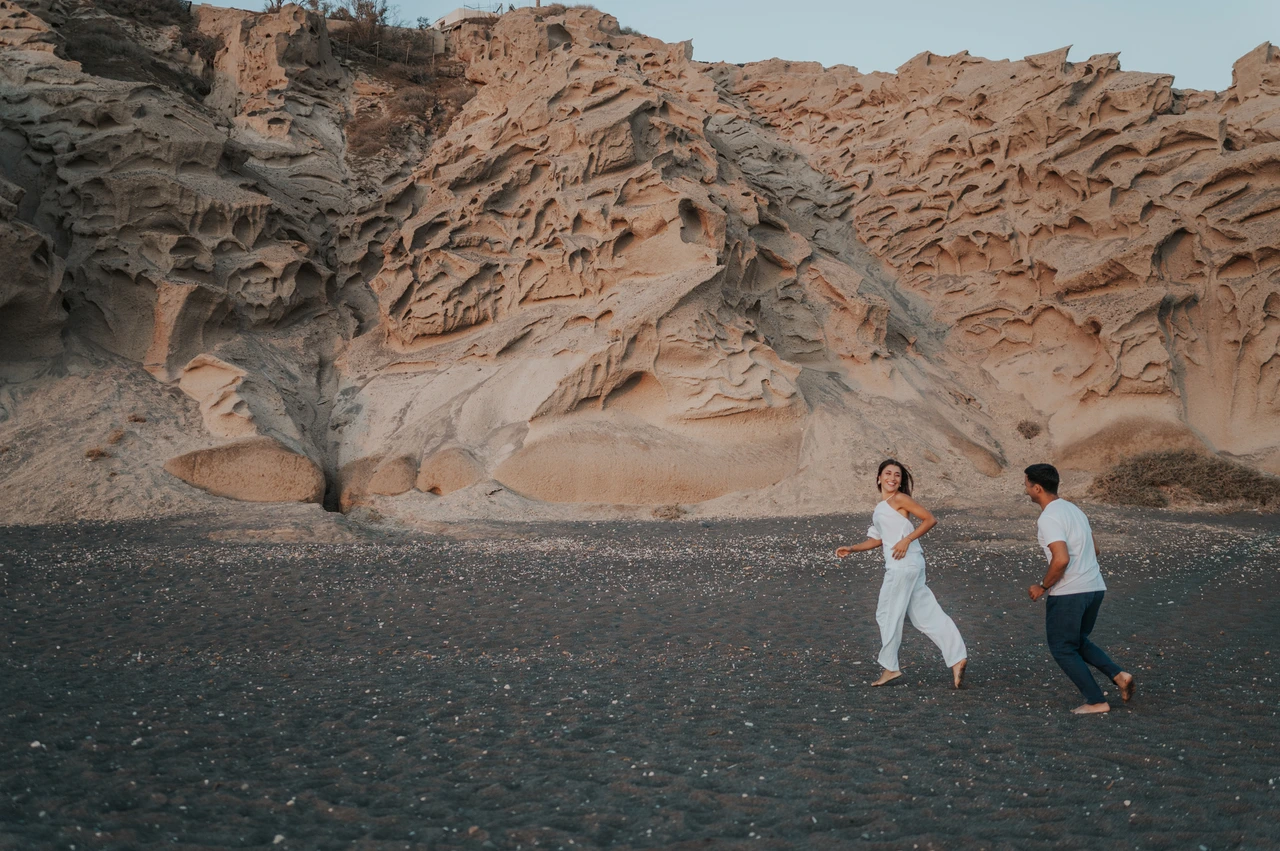 Santorini couple photoshoot: running below Vlychada’s sculpted cliffs Wide view of a couple running hand in hand across dark sand below sculpted rock formations during their Santorini couple photoshoot.