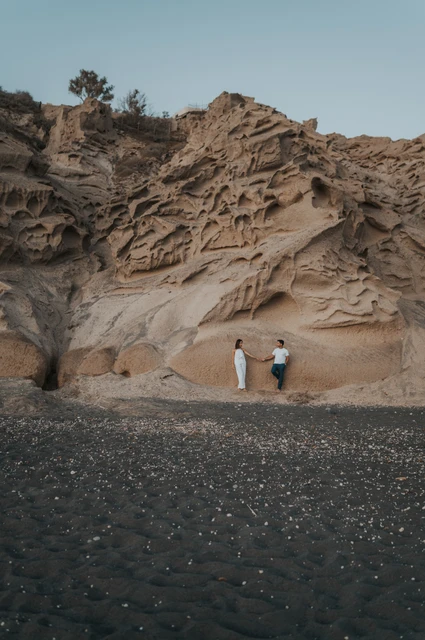 Santorini couple photoshoot: small figures beneath Vlychada’s tall cliffs Wide portrait of a couple standing below tall textured cliffs above the dark beach during their Santorini couple photoshoot.