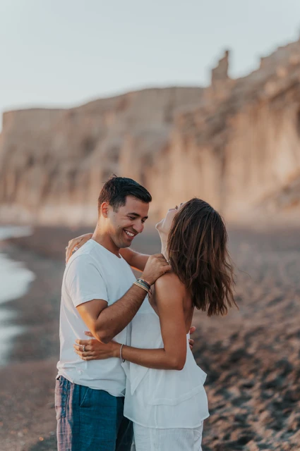 Santorini couple photoshoot: smiles and joined hands beside the cliffs Couple smiling and holding hands with pale sculpted cliffs in the background during their Santorini couple photoshoot.