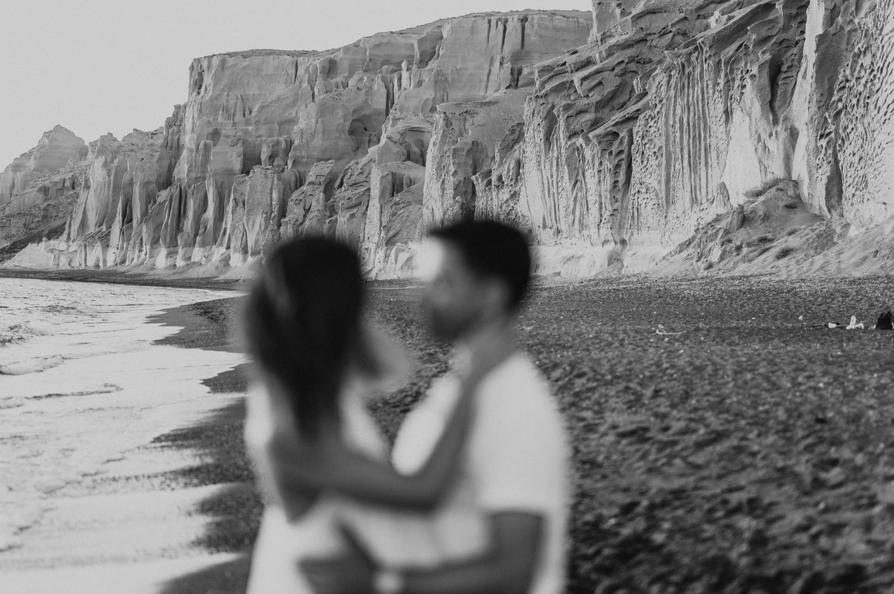 Santorini couple photoshoot: soft-focus black-and-white beach portrait Soft-focus black-and-white portrait of a couple embracing with the beach cliffs behind them during their Santorini couple photoshoot.