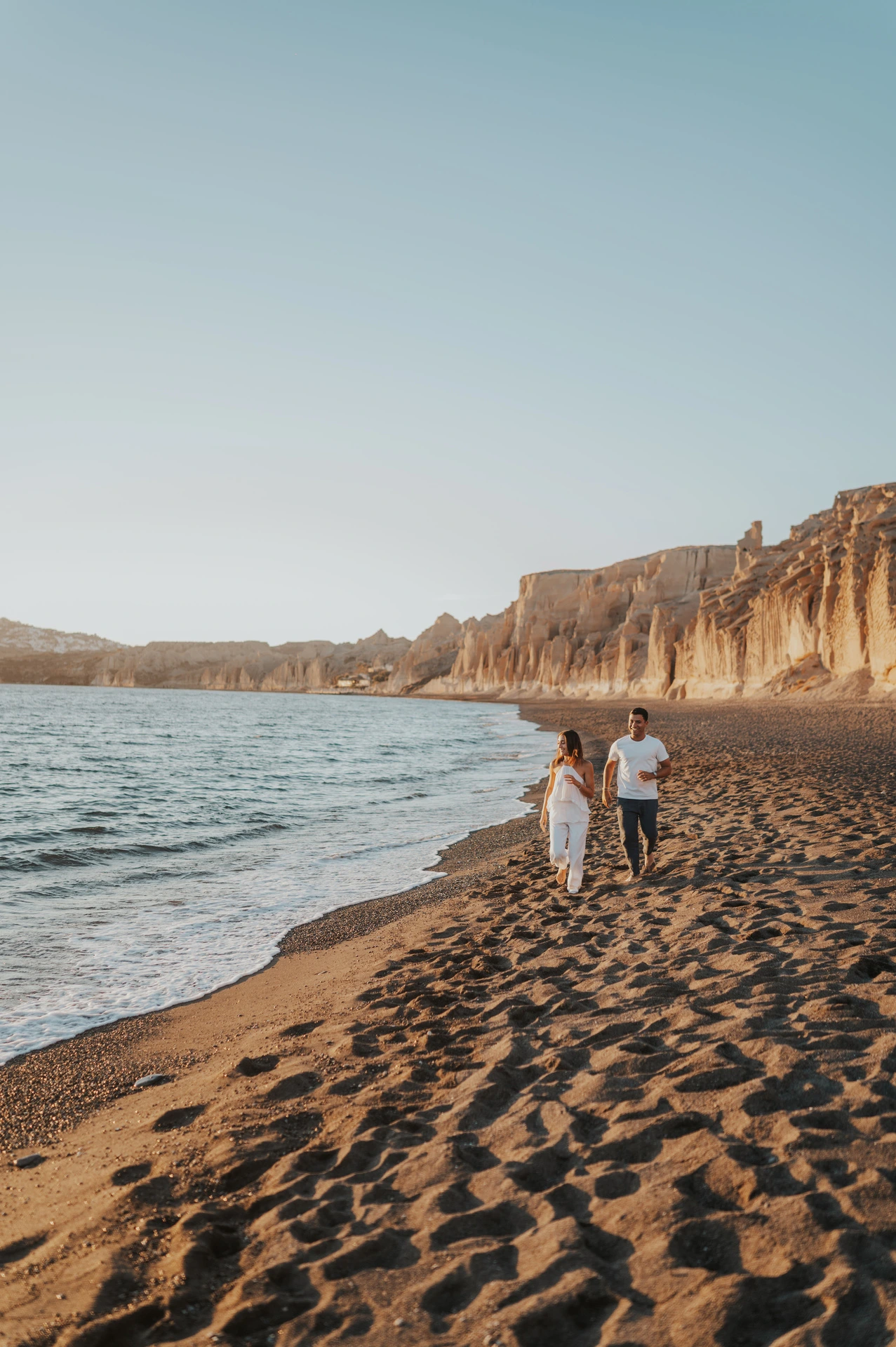 Wide portrait of a couple walking along the shoreline beneath Vlychada’s pale cliffs during their Santorini couple photoshoot.
