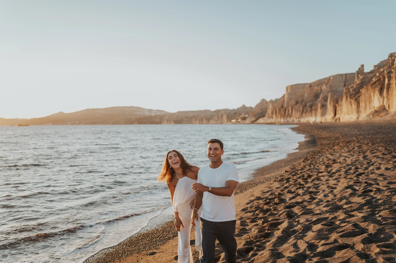 Santorini couple photoshoot: walking beside the water at Vlychada Couple walking towards the camera beside the water with pale cliffs behind them during their Santorini couple photoshoot.
