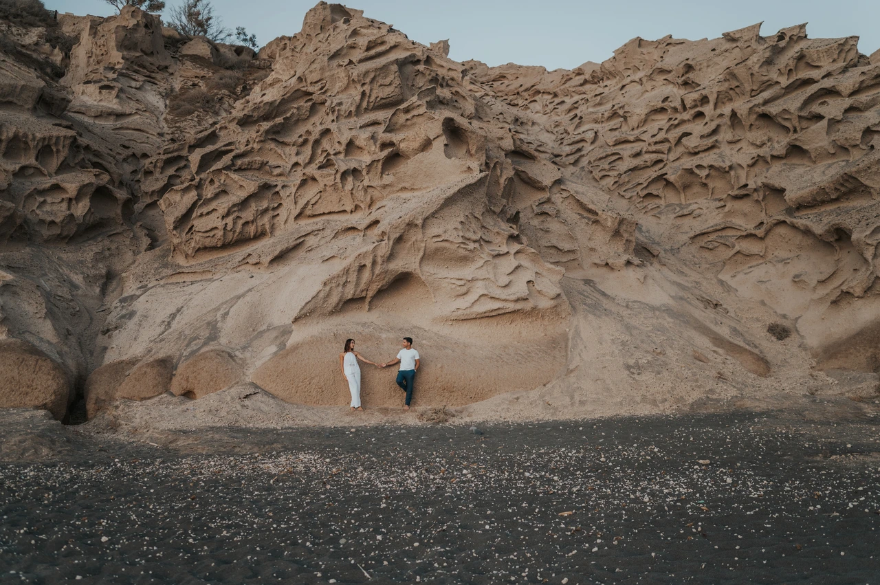 Santorini couple photoshoot: wide portrait beneath sculpted rock Couple standing beneath a broad wall of pale sculpted rock above dark sand during their Santorini couple photoshoot.