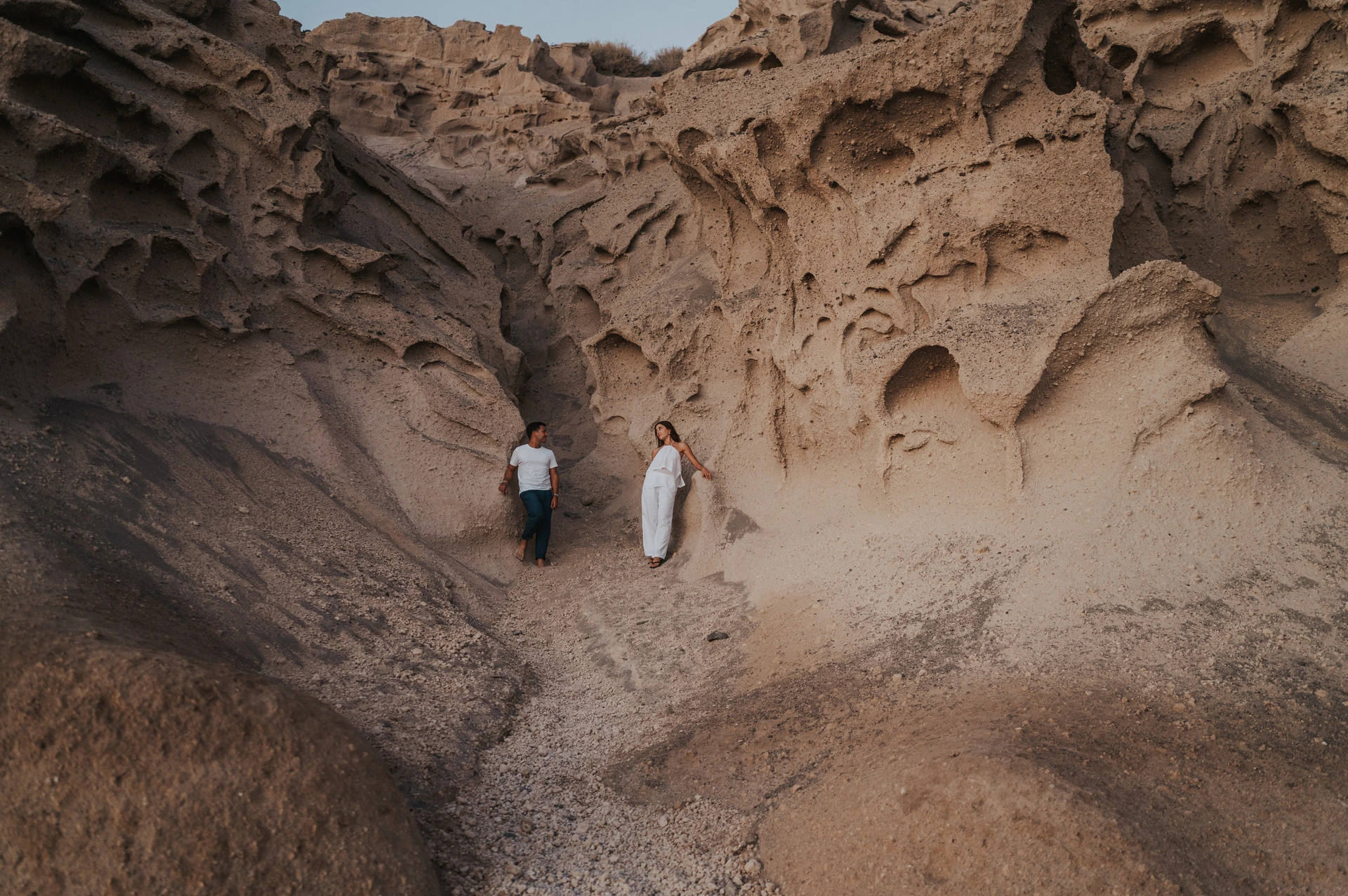 Wide view of a couple standing apart in a pale sculpted rock alcove above the beach during their Santorini couple photoshoot.