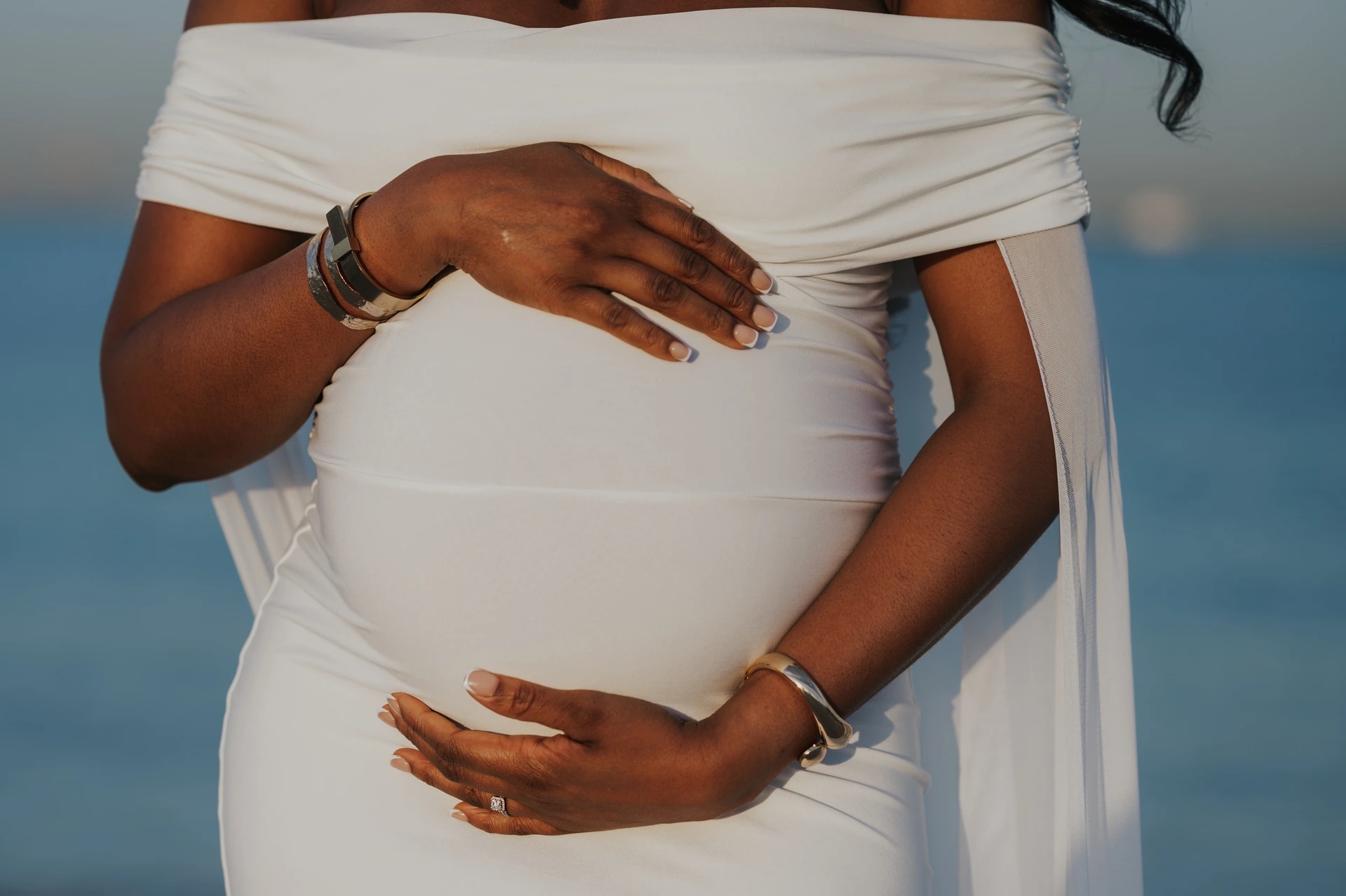 Gros plan d’une femme enceinte en robe blanche tenant son ventre lors d’une séance photo de grossesse sur la plage de Dubaï.