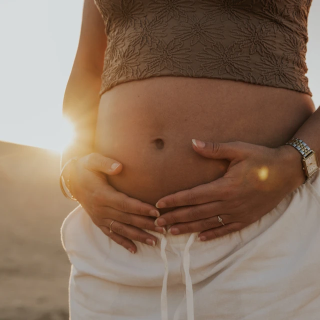 Séance photo grossesse Dubai Gros plan d’un ventre de femme enceinte lors d’une séance photo de grossesse au coucher du soleil dans le désert de Dubaï, avec la lumière du soleil passant derrière les dunes et illuminant son ventre.