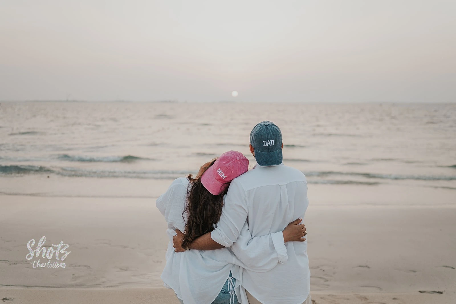Couple enceinte portant des casquettes “mom” et “dad” lors d’une séance photo de grossesse sur la plage de Dubaï au coucher du soleil.