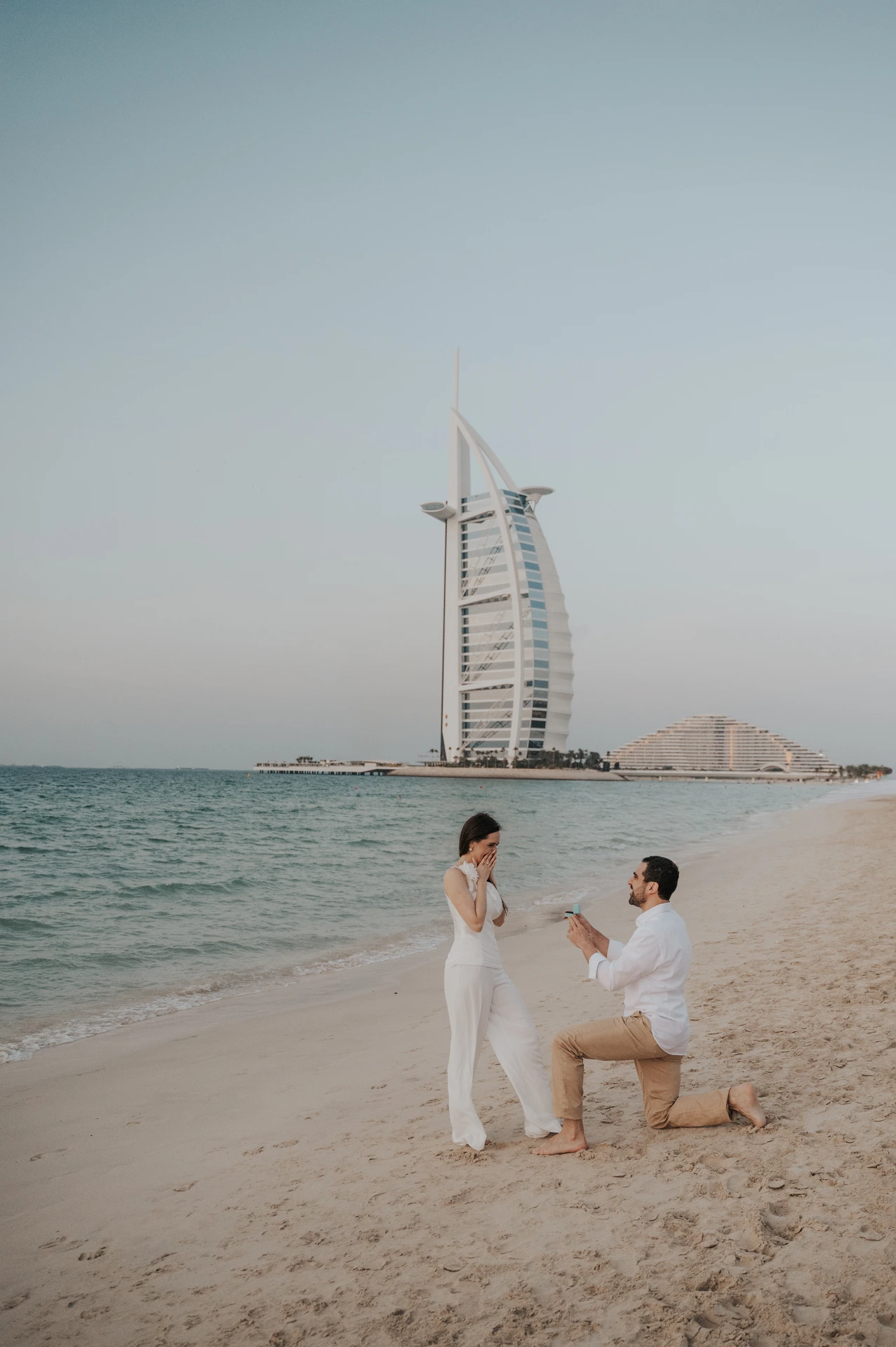 A couple kneels on Dubai beach near Shimmers and shares the ring reveal after a proposal, with Burj Al Arab in view.