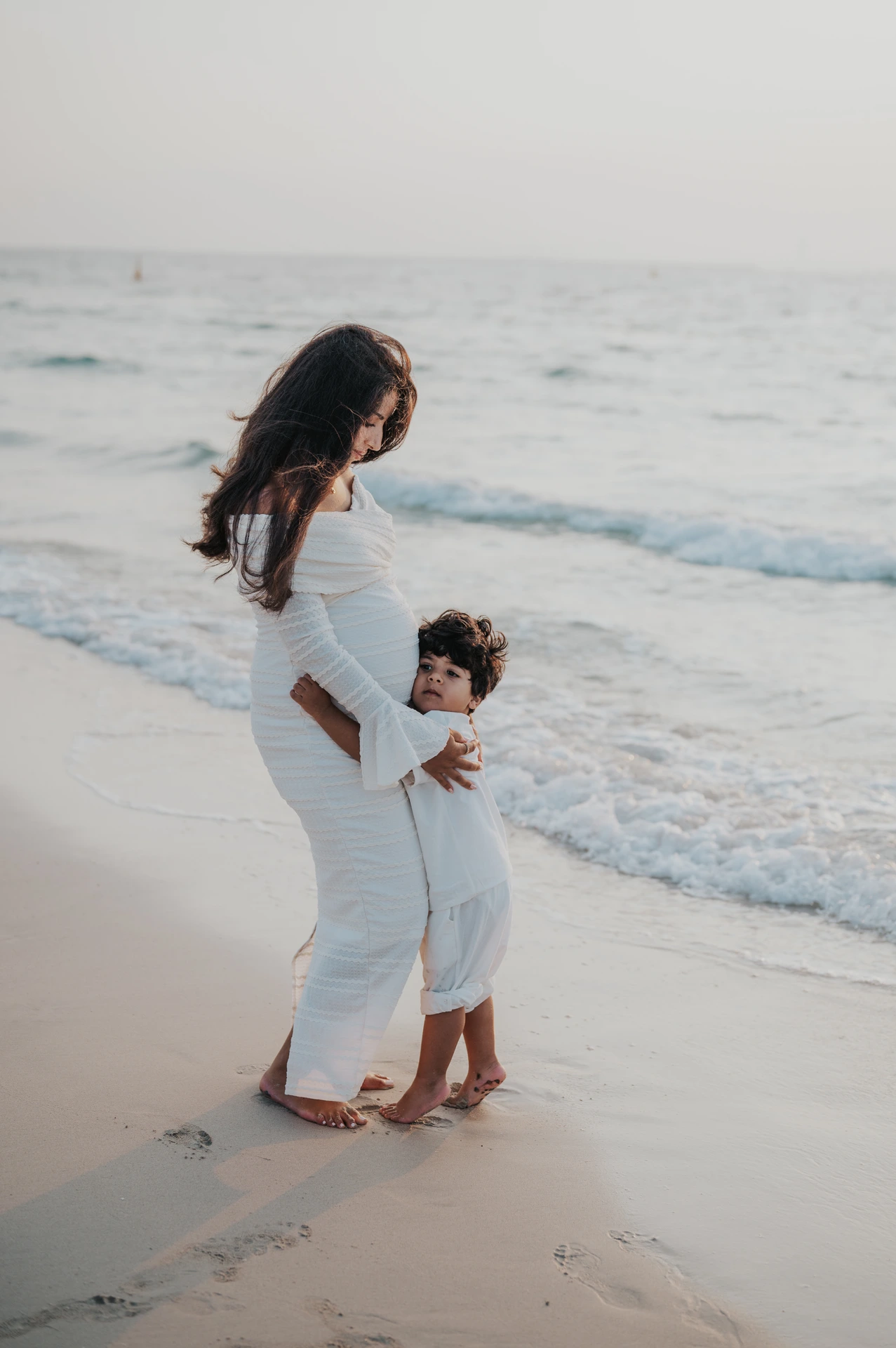 A young child hugs the baby bump while standing with their pregnant mother on Dubai beach during a maternity photoshoot with siblings.