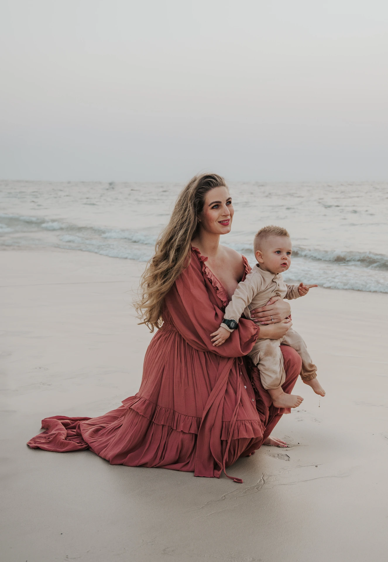 A pregnant mother holds her toddler near the sea in Dubai during a maternity photoshoot with siblings, with soft evening light on the beach.
