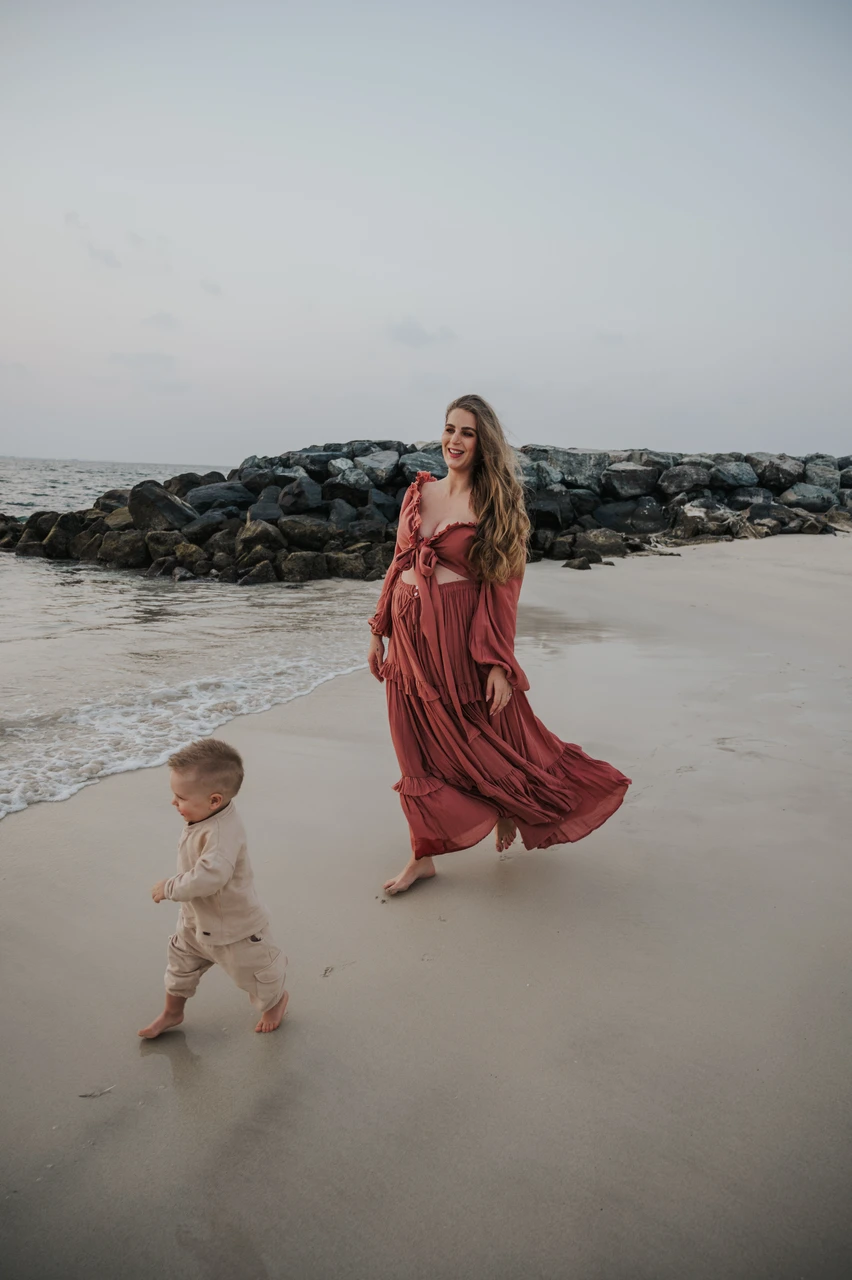Maternity photoshoot with siblings - mother toddler seaside stroll A pregnant mother walks along the beach with her toddler in Dubai during a maternity photoshoot with siblings, framed by shoreline rocks and gentle waves.