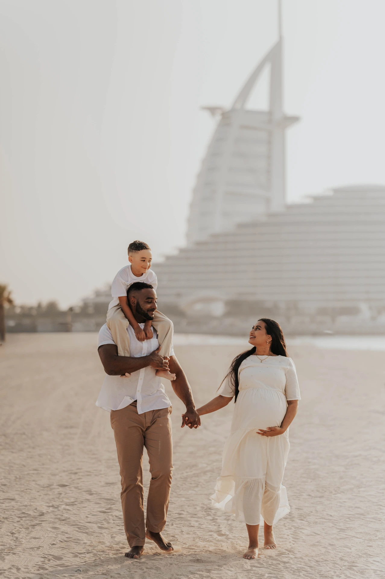 A pregnant mother walks with her partner and child on a Dubai beach during a maternity photoshoot with siblings, with Burj Al Arab softly visible behind them.